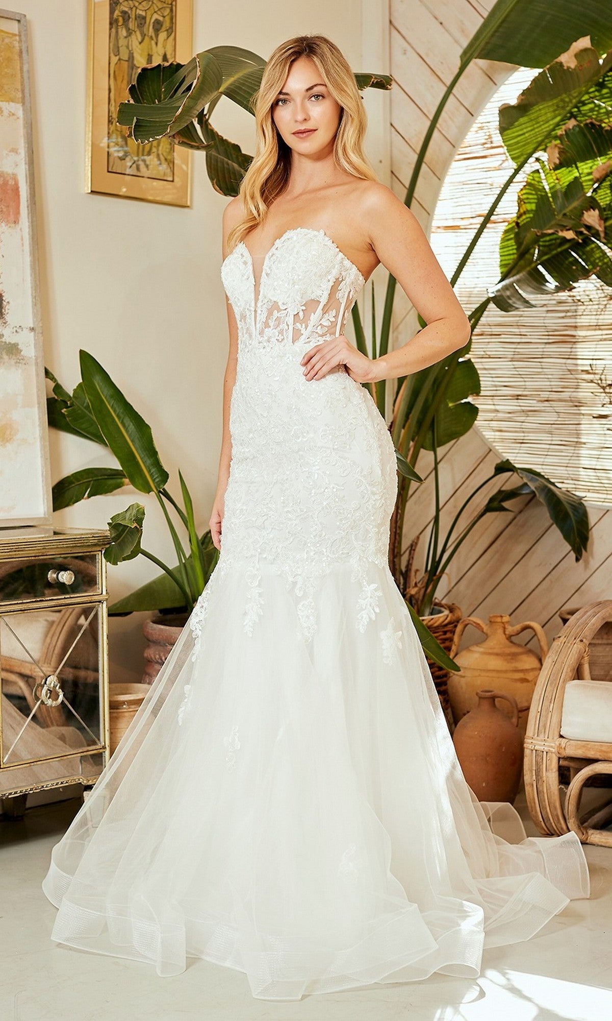 A woman stands indoors wearing the Amelia Couture 7090 long prom dress, featuring a strapless mermaid silhouette with a white lace illusion corset bodice and tulle skirt, surrounded by plants and neutral decor.